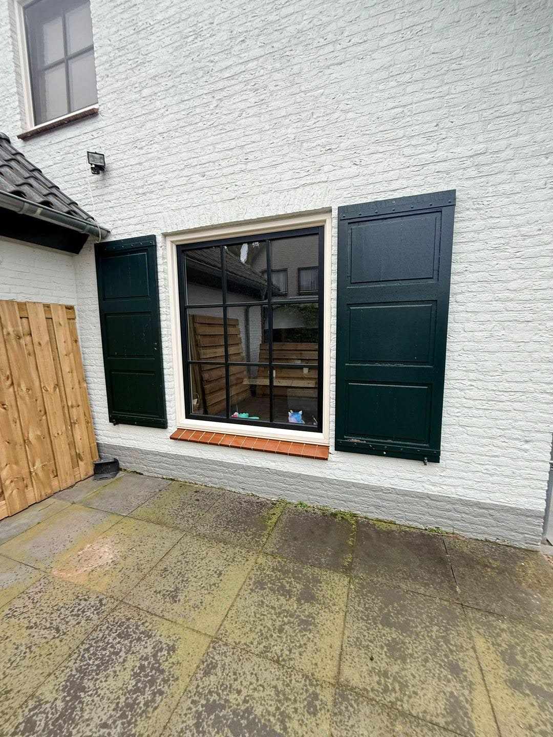 Raam met twee donkere luiken in de witte gevel, naast een houten tuinpoort