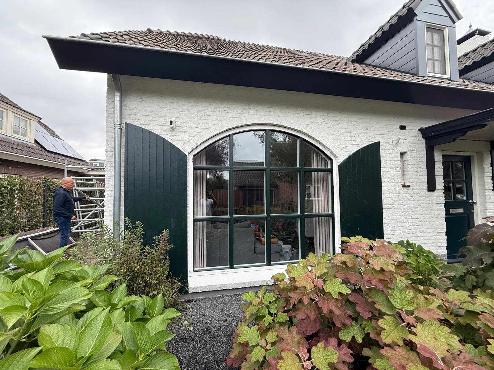 Witte gevel met boogvenster en donkergroene luiken, een schilder op de steiger links