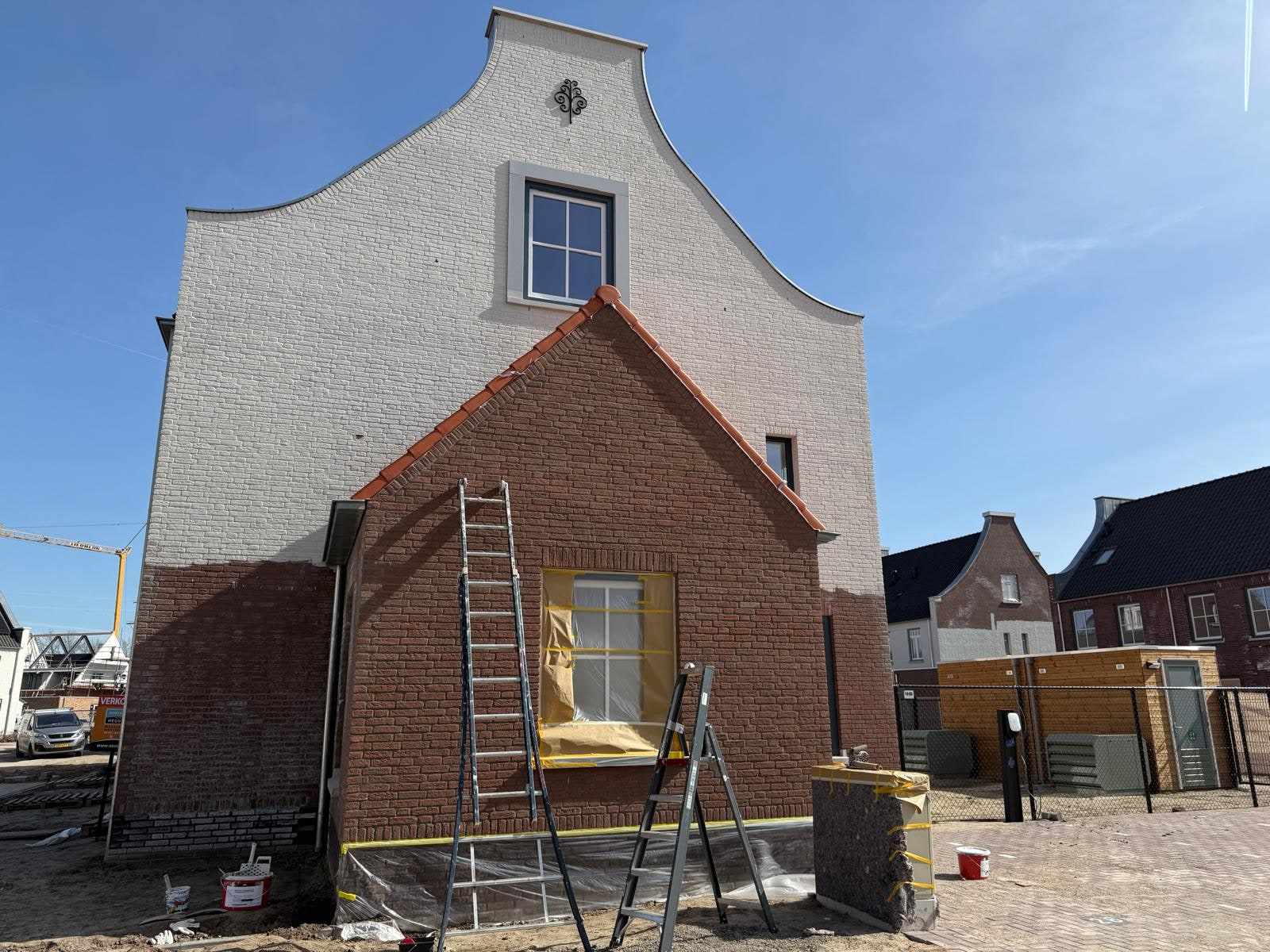 Witte trapgevel boven een nog rode bakstenen aanbouw met ladders ervoor