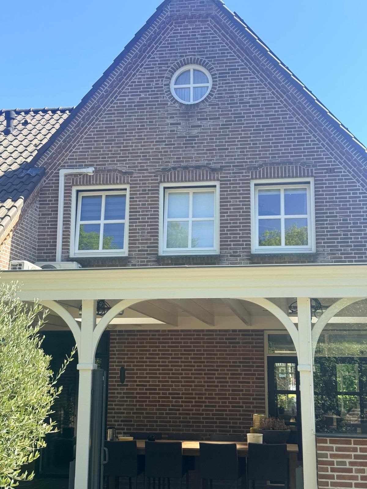 Voorgevel met puntgevel, rond zolderraam en witte kozijnen boven de houten overkapping