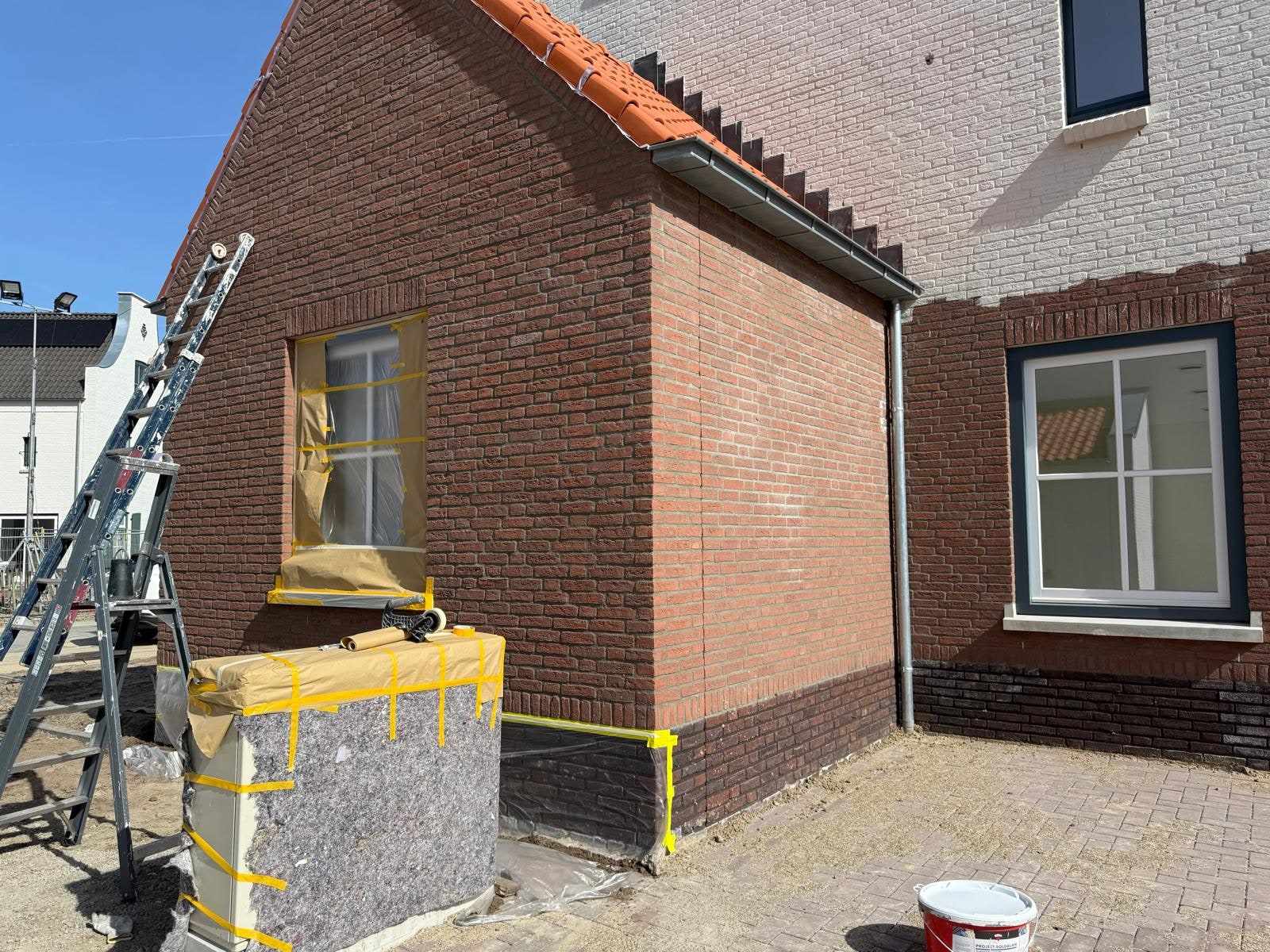 Rode bakstenen zijgevel met afgeplakt raam en ladders, de witte gevel al zichtbaar erachter