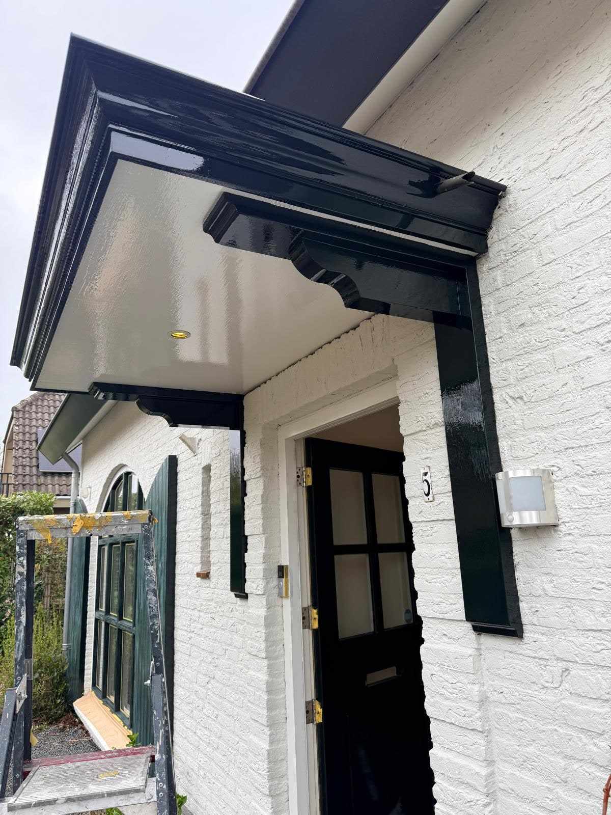 Zwarte luifel met houten consoles boven de entree van de witte woning